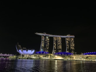 A nighttime view of a modern, iconic hotel with three tower structures connected by a sky terrace. The buildings are illuminated with warm lights, reflecting in a body of water. A uniquely shaped adjacent building features a futuristic design, and the cityscape is subtly lit, creating a vibrant nocturnal atmosphere.