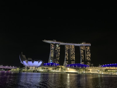 A nighttime view of a modern, iconic hotel with three tower structures connected by a sky terrace. The buildings are illuminated with warm lights, reflecting in a body of water. A uniquely shaped adjacent building features a futuristic design, and the cityscape is subtly lit, creating a vibrant nocturnal atmosphere.