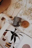 A cozy and aesthetically pleasing arrangement featuring a knitted sweater, a black candle labeled 'HOUSE warming black amber', some delicate dried flowers, and an illustration with abstract and botanical designs. The overall setup exudes warmth and a sense of calm.