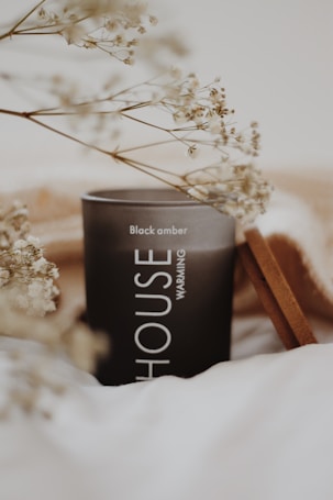 A dark candle labeled 'Black amber' is surrounded by soft, blurry foreground elements of dried white flowers. The scene is set on a soft white and beige background, creating a warm and cozy atmosphere.