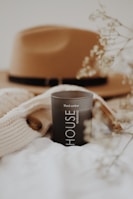 A black candle labeled 'HOUSE WARMING' with 'Black amber' text is positioned on a soft, white fabric surface. A beige knitted sweater with a wooden button is nearby. In the blurred background, a brown felt hat with a black band is visible. Delicate dried flowers are partially in focus, enhancing the soft, cozy aesthetic.