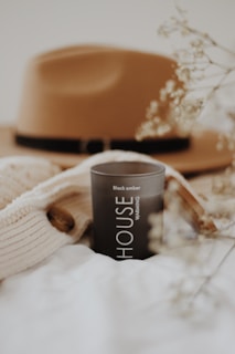 A black candle labeled 'HOUSE WARMING' with 'Black amber' text is positioned on a soft, white fabric surface. A beige knitted sweater with a wooden button is nearby. In the blurred background, a brown felt hat with a black band is visible. Delicate dried flowers are partially in focus, enhancing the soft, cozy aesthetic.