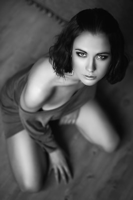 A black and white portrait of a person with short dark hair kneeling on a wooden floor. The focus is on the person's expressive eyes and face. The composition emphasizes an artistic, intimate atmosphere.