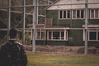 A person is standing in the foreground, observing a rustic green house with multiple windows. The house has a weathered appearance and is surrounded by a metal framework structure. There is a wooden ladder leading up to the roof area. The setting includes grassy land in the foreground and trees in the background.