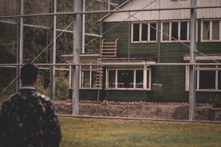 A person is standing in the foreground, observing a rustic green house with multiple windows. The house has a weathered appearance and is surrounded by a metal framework structure. There is a wooden ladder leading up to the roof area. The setting includes grassy land in the foreground and trees in the background.