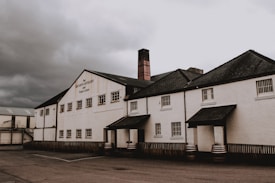 The image portrays a distillery building with a white facade and multiple small windows. The roof is dark and sloped, with two small porch structures at the entrances. A wooden fence runs along the base of the building, and several barrels are visible near the entrance. The sky is overcast with dark, heavy clouds.