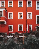 A charming outdoor caf&eacute; surrounded by greenery is set against a vibrant red building with wooden-framed windows. There are several tables and wicker chairs neatly arranged on a cobblestone patio with red umbrellas closed beside them. The windows display various potted plants, adding to the cozy and inviting atmosphere.