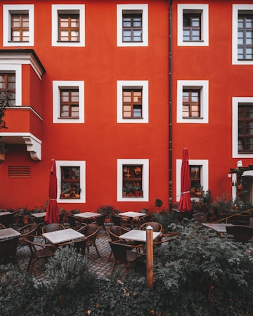 A charming outdoor caf&eacute; surrounded by greenery is set against a vibrant red building with wooden-framed windows. There are several tables and wicker chairs neatly arranged on a cobblestone patio with red umbrellas closed beside them. The windows display various potted plants, adding to the cozy and inviting atmosphere.