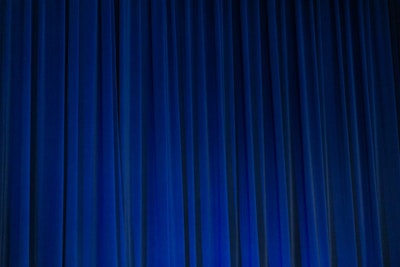A set of vertical, evenly spaced blue curtains appear to be drawn across a stage or large window. The fabric has a dense texture and the lighting creates subtle shadows and highlights.