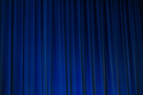 A set of vertical, evenly spaced blue curtains appear to be drawn across a stage or large window. The fabric has a dense texture and the lighting creates subtle shadows and highlights.