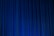 A set of vertical, evenly spaced blue curtains appear to be drawn across a stage or large window. The fabric has a dense texture and the lighting creates subtle shadows and highlights.