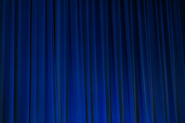 A set of vertical, evenly spaced blue curtains appear to be drawn across a stage or large window. The fabric has a dense texture and the lighting creates subtle shadows and highlights.