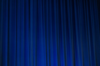 A set of vertical, evenly spaced blue curtains appear to be drawn across a stage or large window. The fabric has a dense texture and the lighting creates subtle shadows and highlights.