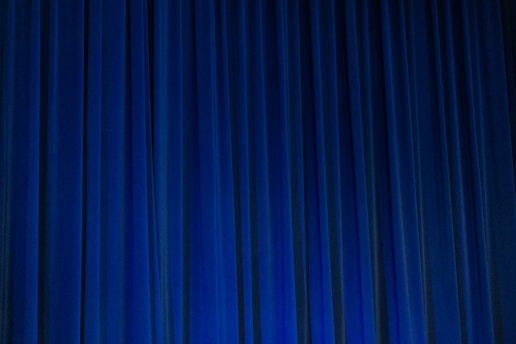 A set of vertical, evenly spaced blue curtains appear to be drawn across a stage or large window. The fabric has a dense texture and the lighting creates subtle shadows and highlights.