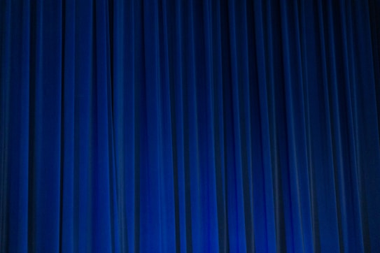 A set of vertical, evenly spaced blue curtains appear to be drawn across a stage or large window. The fabric has a dense texture and the lighting creates subtle shadows and highlights.