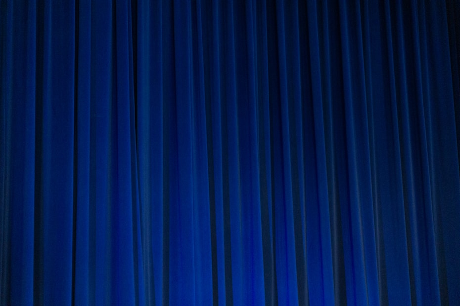 A set of vertical, evenly spaced blue curtains appear to be drawn across a stage or large window. The fabric has a dense texture and the lighting creates subtle shadows and highlights.
