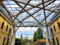A modern glass atrium with a geometric steel frame supporting the roof structure. The walls are lined with beige-colored buildings, and large green trees can be seen through the expansive windows. The ceiling features a network of light fixtures and the sky is partially visible with some clouds.