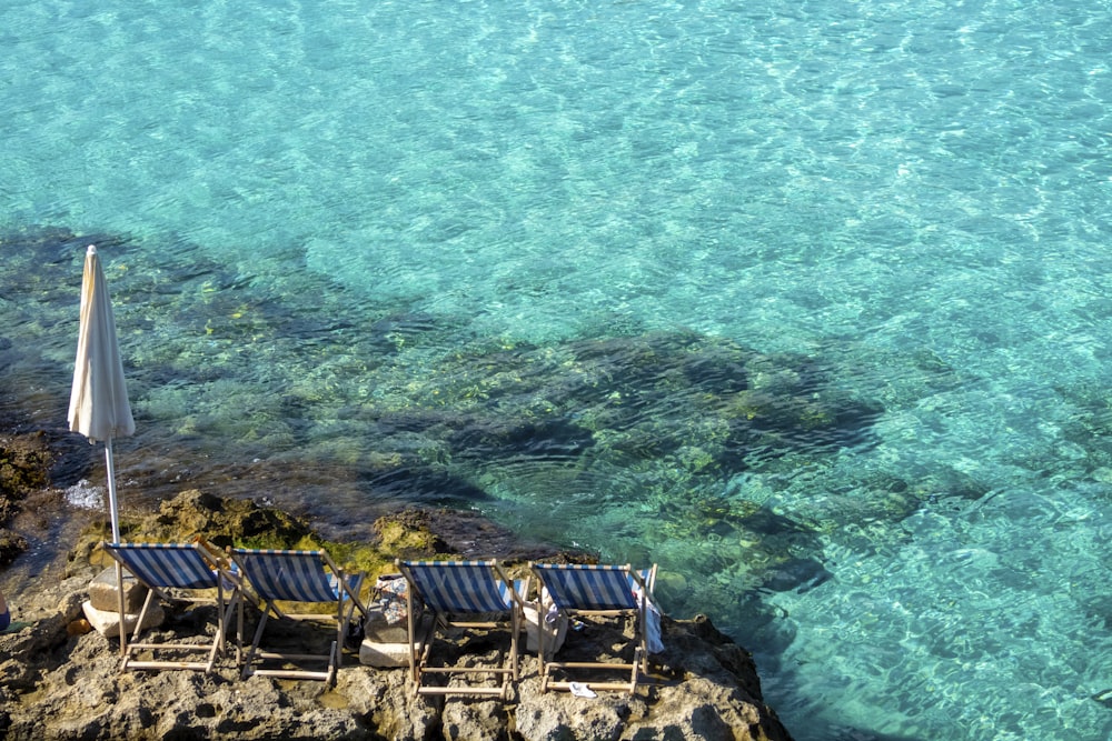 Quattro lettini a strisce bianche e blu sulla roccia con ombrellone vicino a uno specchio d'acqua limpido