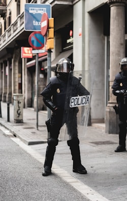 A police officer dressed in riot gear stands on a city street holding a transparent shield marked 'Policia'. The background features urban elements such as street signs, including a bus and taxi sign, and a 'no entry' sign, as well as buildings with columns.