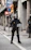 A police officer dressed in riot gear stands on a city street holding a transparent shield marked 'Policia'. The background features urban elements such as street signs, including a bus and taxi sign, and a 'no entry' sign, as well as buildings with columns.