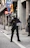 A police officer dressed in riot gear stands on a city street holding a transparent shield marked 'Policia'. The background features urban elements such as street signs, including a bus and taxi sign, and a 'no entry' sign, as well as buildings with columns.