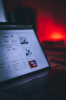 A laptop displaying a dashboard with various graphs, charts, and numeric data. The screen is partially visible, showcasing sections with line graphs, financial figures, and small images. The background is blurred with a prominent red light creating a warm ambiance.
