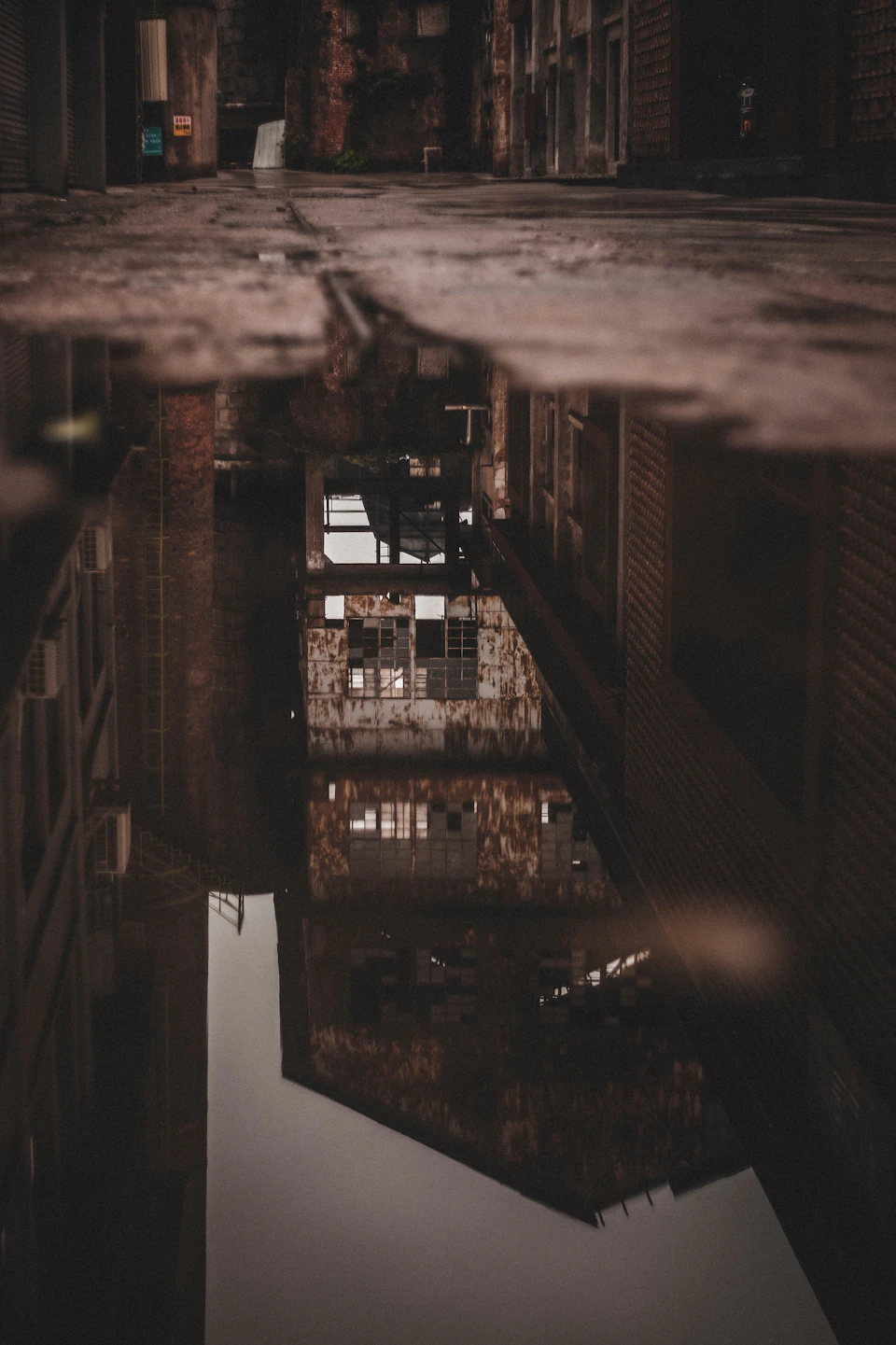 The image shows a reflection of an old brick building, possibly a warehouse or factory, in a puddle on the ground. The building's facade is visible through the watery surface, creating a mirror-like effect that adds depth to the scene. The perspective of the photo is taken from below, looking up at the building and its reflection, giving the viewer a sense of being part of the scene.