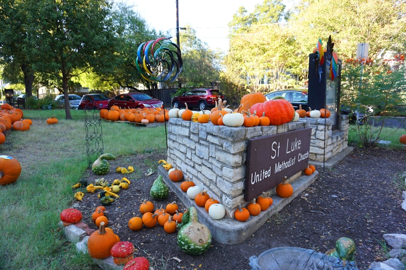 A church sign surrounded by numerous pumpkins of varying sizes and colors, including orange, white, and green gourds, set in a grassy area. Trees and parked cars are in the background, and the sign reads 'St Luke United Methodist Church'. There is also a colorful sculpture of concentric circles nearby.