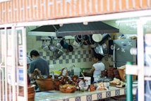 A busy kitchen setting with two people working. Pots and pans hang on the tiled wall. The countertop is filled with various ceramic dishes and food items. A refrigerator is visible in the background alongside more kitchen equipment.