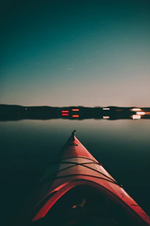 A red kayak is seen from the perspective of someone sitting inside, pointed towards a calm, reflective body of water during twilight. The horizon is lined with faint silhouettes of land, and some distant lights create small reflections on the water surface.