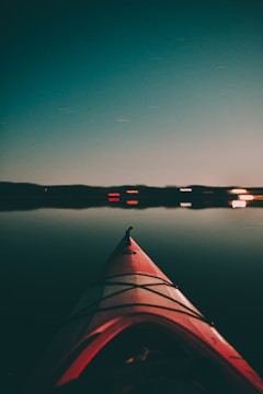 A red kayak is seen from the perspective of someone sitting inside, pointed towards a calm, reflective body of water during twilight. The horizon is lined with faint silhouettes of land, and some distant lights create small reflections on the water surface.