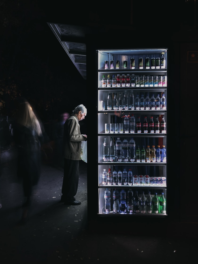 A man stands in front of a well-lit vending machine stocked with various beverages. The scene is set in a dimly lit environment, creating a stark contrast between the bright vending machine and the dark surroundings.