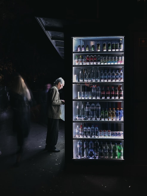 A man stands in front of a well-lit vending machine stocked with various beverages. The scene is set in a dimly lit environment, creating a stark contrast between the bright vending machine and the dark surroundings.