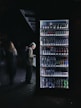 A man stands in front of a well-lit vending machine stocked with various beverages. The scene is set in a dimly lit environment, creating a stark contrast between the bright vending machine and the dark surroundings.