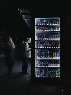 A man stands in front of a well-lit vending machine stocked with various beverages. The scene is set in a dimly lit environment, creating a stark contrast between the bright vending machine and the dark surroundings.