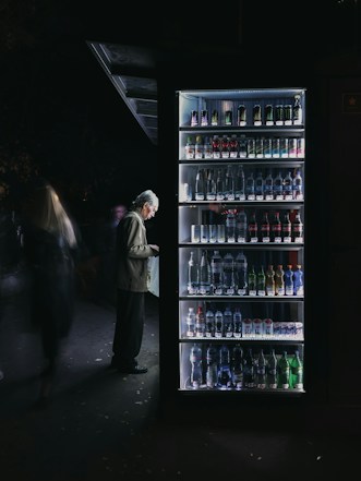 A man stands in front of a well-lit vending machine stocked with various beverages. The scene is set in a dimly lit environment, creating a stark contrast between the bright vending machine and the dark surroundings.