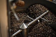 A close-up view of freshly roasted coffee beans inside a coffee roasting machine. The machine's interior has metallic components, with an agitator arm stirring the coffee beans. The beans appear dark brown, indicating a rich roast.