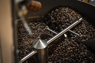 A close-up view of freshly roasted coffee beans inside a coffee roasting machine. The machine's interior has metallic components, with an agitator arm stirring the coffee beans. The beans appear dark brown, indicating a rich roast.