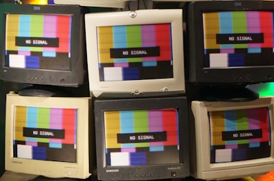 A collection of vintage CRT monitors displaying a 'No Signal' message over a multicolored test pattern screen. The screens show a mix of red, green, blue, yellow, and white stripes. The monitors are arranged closely together, showcasing different brands such as IBM and Samsung.