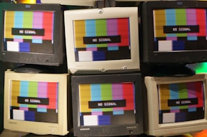 A collection of vintage CRT monitors displaying a 'No Signal' message over a multicolored test pattern screen. The screens show a mix of red, green, blue, yellow, and white stripes. The monitors are arranged closely together, showcasing different brands such as IBM and Samsung.
