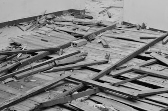 Broken wooden floorboards and scattered debris cover the ground in an indoor space. The floor appears damaged with displaced panels and exposed nails, giving an impression of neglect or abandonment.