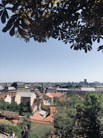 A scenic cityscape with red-roofed buildings spread out under a clear blue sky. In the foreground, leafy branches frame the top of the image, and a pair of people are walking along a pathway. A sign indicating a food bar and caf&eacute; is visible. The city extends into the distance, with various architectural styles and a few taller structures on the horizon.