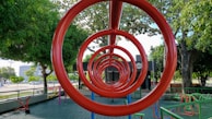 A playground features red circular climbing bars forming a tunnel-like perspective. Surrounding the structure are various playground equipment and large, leafy trees providing shade. The ground is covered with green tiles and there are fences bordering the area, with a road and buildings visible in the background.