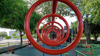 A playground features red circular climbing bars forming a tunnel-like perspective. Surrounding the structure are various playground equipment and large, leafy trees providing shade. The ground is covered with green tiles and there are fences bordering the area, with a road and buildings visible in the background.