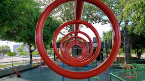A playground features red circular climbing bars forming a tunnel-like perspective. Surrounding the structure are various playground equipment and large, leafy trees providing shade. The ground is covered with green tiles and there are fences bordering the area, with a road and buildings visible in the background.