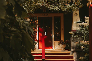 A warm, inviting home entrance bathed in soft natural light with a welcoming red door framed by lush green plants.