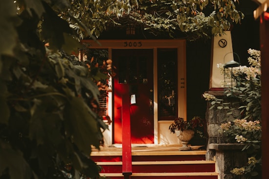 A warmly lit entrance to a house with a vibrant red door and a stone facade entryway. The stoop is decorated with potted plants, and the area is surrounded by lush greenery and overhanging branches that create a cozy, shaded atmosphere.