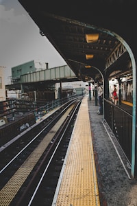 An urban train station platform with metal overhead structures and rail tracks running through. The platform features a yellow safety line on the ground and is partially covered by a dark overhead awning. Industrial buildings and a graffiti-laden wall are visible in the background. A person stands on the platform near a train.