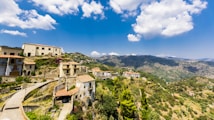 A picturesque hilltop village with rustic buildings nestled among lush greenery. Rolling mountains extend into the background under a bright, blue sky dotted with fluffy white clouds. The tightly clustered houses have terracotta roofs and stone walls.