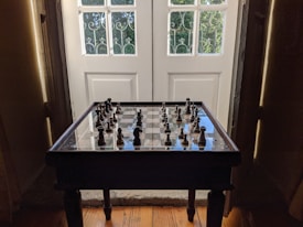 A wooden chessboard with chess pieces set up sits on a small table in front of a large, white, paneled door. The door has square glass panels framed by metal bars, through which greenery can be seen. Warm light filters through, casting shadows and highlighting the chess pieces.